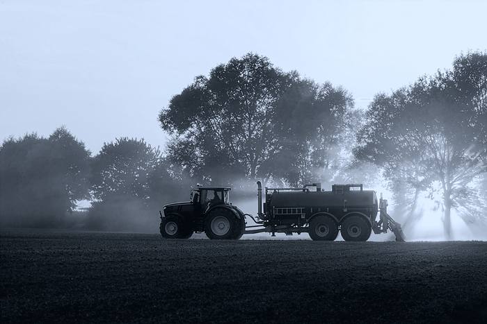 A tractor drawing a tanker across a field at dusk, spray drifting behind it.