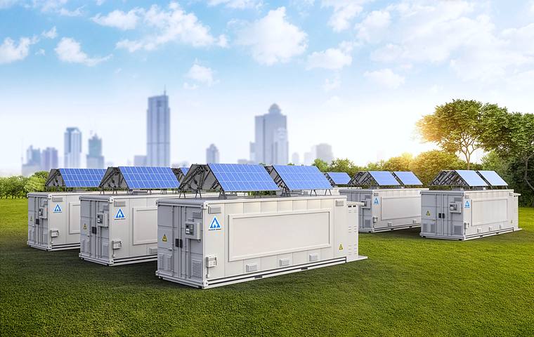 A row of containerised battery energy storage units with solar arrays mounted above them, on the edge of a city.