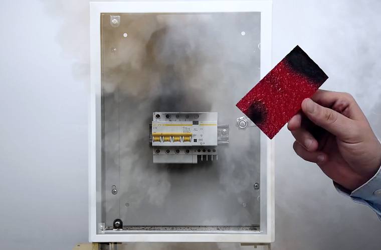 A distribution board filled with smoke after a fire inside it, with a scorched TITAN pad held up beside the open enclosure.
